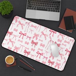 May include: A white desk mat with a pink watercolor bow pattern. The mat is designed for use with a computer mouse and keyboard.