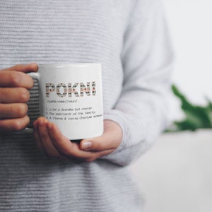 May include: White ceramic mug with the word "POKNI" in a decorative pattern. Below the word is the definition: "Like a Grandma but cooler. The matriarch of the family. A fierce & loving Choctaw woman."