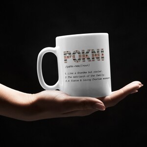 May include: White ceramic mug with the word "POKNI" in a decorative pattern. The mug also includes a definition: "Like a Grandma but cooler," "The matriarch of the family," and "A fierce & loving Choctaw woman."