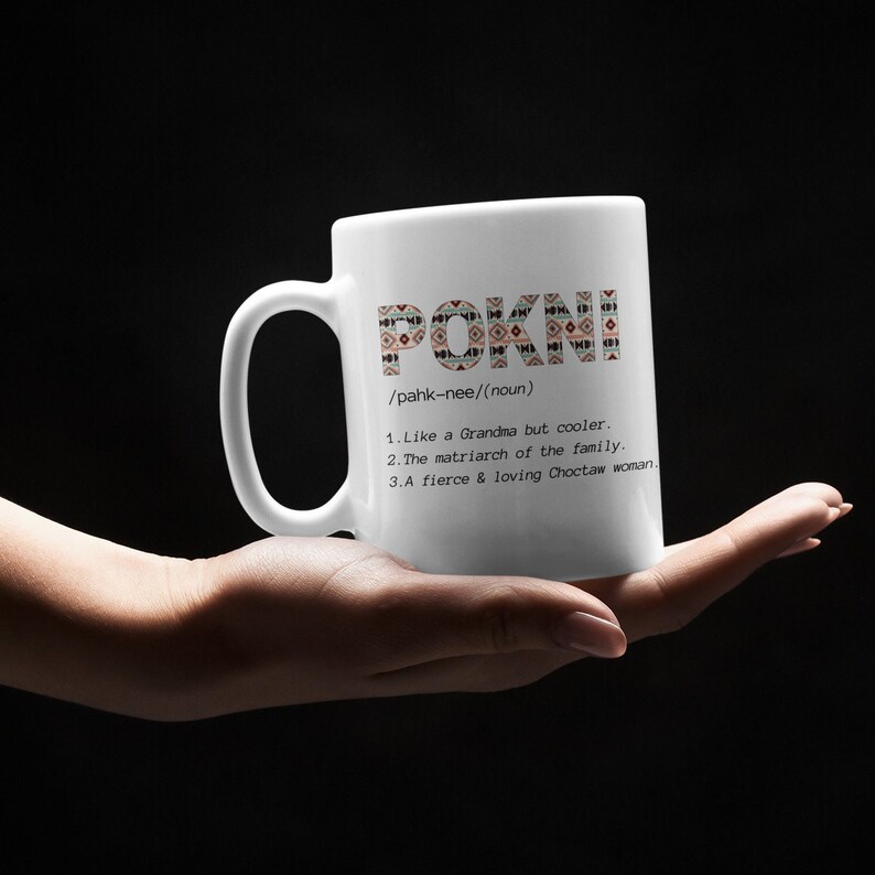 May include: White ceramic mug with the word "POKNI" in a decorative pattern. The mug also includes a definition: "Like a Grandma but cooler," "The matriarch of the family," and "A fierce & loving Choctaw woman."