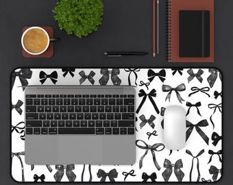 May include: A white desk mat with a black and white watercolor bow pattern. A laptop computer, a wireless mouse, and a cup of coffee are on the desk mat.