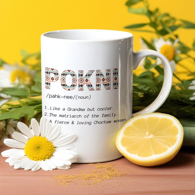 May include: White ceramic mug with the word "POKNI" in a patterned design. Below the word are definitions: "Like a Grandma but cooler," "The matriarch of the family," and "A fierce & loving Choctaw woman."
