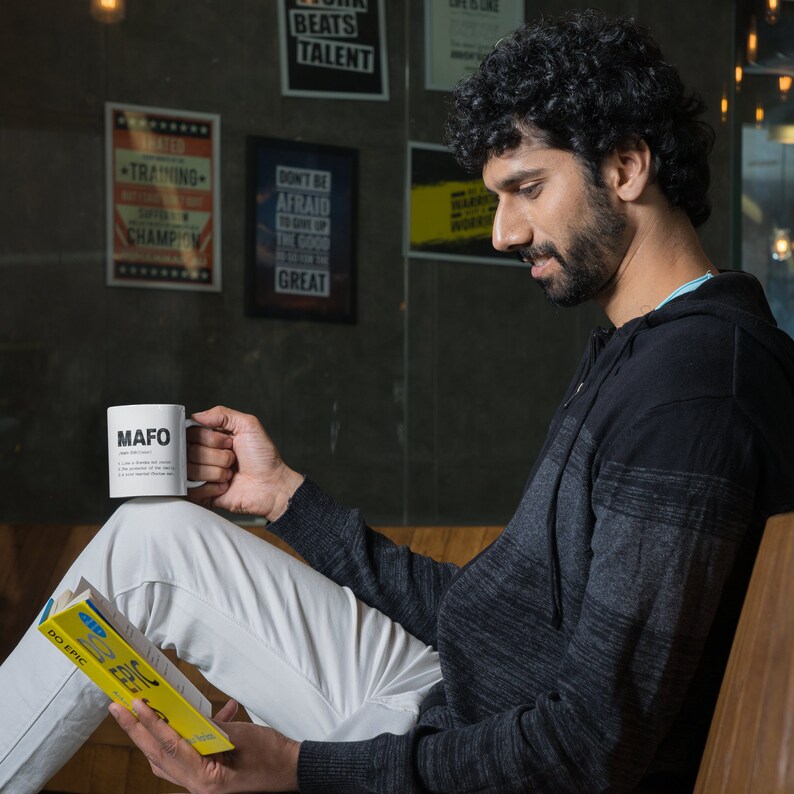 May include: White ceramic mug with the acronym "MAFO" printed in black, held by a person. The mug also has a definition printed below the acronym. The person is reading a book with the words "DO EPIC" on the cover.