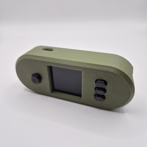 May include: An olive green handheld device with a small black screen and three black buttons on the right. A single black button is on the left. The device has rounded edges and a small hole on the top.