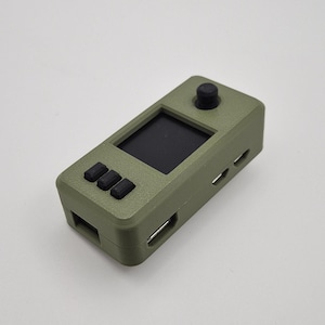 May include: Olive green handheld gaming device with a small black screen, three black buttons, and a black joystick. The device has a rectangular shape with rounded edges and several ports.