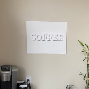 Op de afbeelding: Een wit bord met het woord "COFFEE" in reliëf hangt aan een muur boven een zwart aanrecht met een koffiezetapparaat, twee mokken en een kleine gootsteen. Een potplant met bamboe staat op het aanrecht.