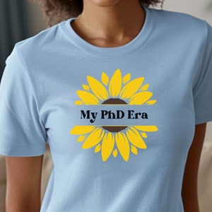 May include: A light blue t-shirt with a yellow sunflower graphic. The sunflower has a brown center and a black banner that reads "My PhD Era".