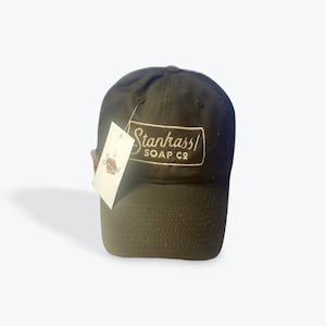 Könnte beinhalten: Dunkelgrüne Baseballkappe mit cremefarbenem, gesticktem Text "Stankass! SOAP CO". Ein weißes Etikett mit dem Logo "Stankass! SOAP CO" ist an der Kappe befestigt.