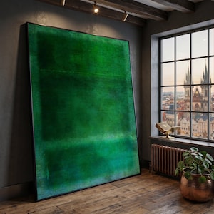 May include: Large framed abstract artwork in shades of green. The painting is set against a dark grey wall in a room with a window offering a view of a city. The frame is black.