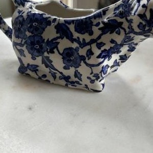 May include: A white ceramic creamer in the shape of a cow with a blue and white floral pattern.