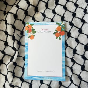 May include: A rectangular notepad with a watercolour design featuring oranges and leaves. The notepad has a blue border and the text "Notes Yaffa, Palestine" and "Falasteen Favorites". The notepad is on a black and white patterned cloth.