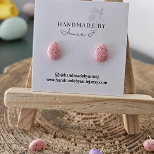 May include: A pair of pink speckled egg-shaped stud earrings on a white card with the text "HANDMADE BY Amie J" and the social media handle "@handmadebyamiej" and website "www.handmadebyamiej.etsy.com".