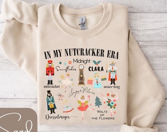 Peut inclure: Un sweat-shirt beige avec un imprimé graphique sur le thème de Casse-Noisette. L'impression comprend le texte "In My Nutcracker Era" et présente des illustrations de personnages et d'objets du ballet Casse-Noisette, tels qu'un soldat casse-noisette, des flocons de neige, une horloge, Clara, un roi des souris, Sugar Plum et une valse des fleurs.