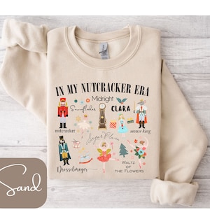 May include: A beige sweatshirt with a graphic print of a Nutcracker theme. The print includes the text "In My Nutcracker Era" and features illustrations of characters and objects from the Nutcracker ballet, such as a nutcracker soldier, snowflakes, a clock, Clara, a mouse king, Sugar Plum, and a waltz of the flowers.