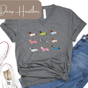 May include: Deep heather grey unisex V-neck t-shirt with a pattern of dachshunds in different outfits. The shirt is styled with blue jeans and white trainers. The text "Deep Heather" and "Unisex V-Neck" are displayed.