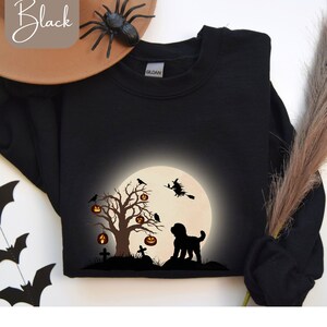 Op de afbeelding: Zwarte Halloween-trui met een afbeelding van een volle maan, een boom met pompoenlantaarns, een heks op een bezem en een honden silhouet. Het woord "Black" staat in een scriptletter in de linkerbovenhoek.