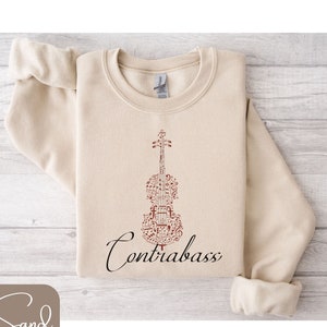 Puede incluir: Una sudadera beige con un contrabajo marrón hecho de notas musicales. El texto "Contrabass" está escrito en cursiva negra debajo del instrumento.