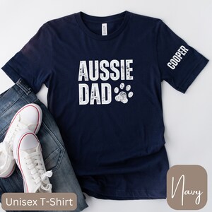 May include: Navy blue unisex t-shirt with the text "Aussie Dad" and a paw print in white. The text "Cooper" is printed on the sleeve.