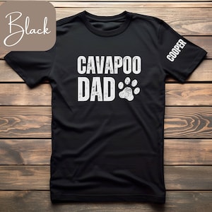 Può includere: Maglietta nera con testo bianco che recita "Cavapoo Dad" e un'impronta di zampa. La manica destra ha il nome "Cooper" stampato.