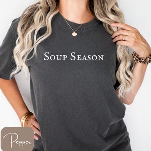 Puede incluir: Camiseta gris oscuro con la frase "Soup Season" en blanco. La camiseta tiene mangas cortas y un corte holgado. La palabra "Pepper" est&aacute; en un cuadrado marr&oacute;n claro en la esquina inferior izquierda.