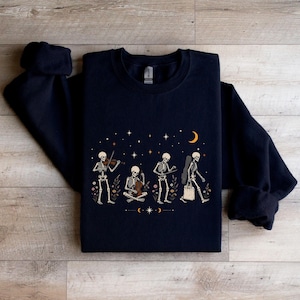 May include: A dark navy blue crewneck sweatshirt featuring a graphic print of four skeletons. One plays a violin, one sits playing a cello, one plays a guitar, and one walks carrying a tote bag. The design includes small flowers, stars, and a crescent moon.