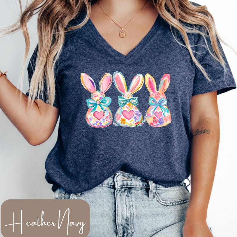 Op de afbeelding: Marineblauwe V-hals T-shirt met een aquarelontwerp van drie paashazen. Elke haas heeft roze oren, een blauwe strik en een hart. De konijnen zijn versierd met kleurrijke bloemmotieven.