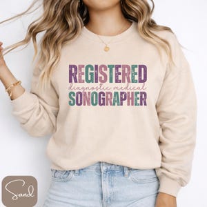 Könnte beinhalten: Beiges Sweatshirt mit dem Text "REGISTERED diagnostic medical SONOGRAPHER" in Lila, Türkis und Grün. Das Sweatshirt hat lange Ärmel und einen Rundhalsausschnitt. Der Text ist in einer serifenlosen Schriftart.