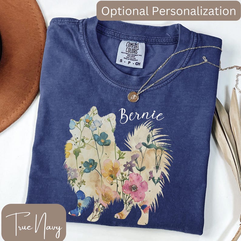May include: A navy blue t-shirt featuring a floral Pomeranian design with the name "Bernie" in white script. The shirt has a crew neck and short sleeves. A gold necklace with a pendant is draped across the shirt. The words "True Navy" are at the bottom.