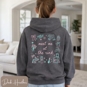 May include: Dark heather gray hoodie with a graphic on the back that says "meet me at the rink" in a whimsical font. The graphic includes ice skates, flowers, and other winter-themed illustrations in teal and pink.