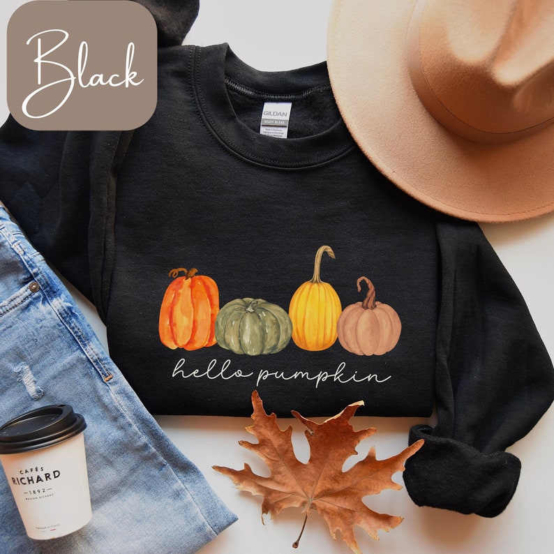 May include: Black sweatshirt with a watercolor design of four pumpkins in orange, green, and yellow. The words "hello pumpkin" are written in white script below the pumpkins. A brown hat and jeans are in the background.