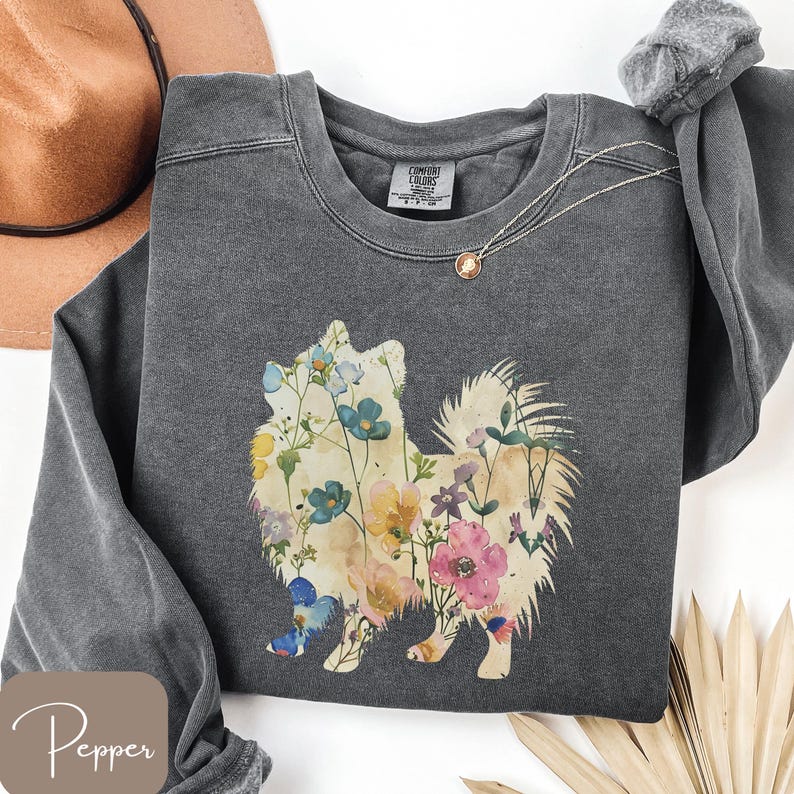 May include: A dark gray sweatshirt featuring a floral dog silhouette. The dog is filled with colorful flowers in shades of blue, pink, yellow, and white. The sweatshirt has a crew neck and long sleeves. A gold necklace with a pendant is draped on the sweatshirt.