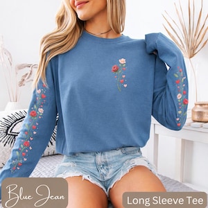 Puede incluir: Una camiseta de manga larga azul vaquero con bordado floral. La camisa presenta un diseño de rosas rojas y rosas, corazones y vegetación en la manga izquierda y el pecho. Las palabras "Blue Jean" están impresas en la parte inferior izquierda.