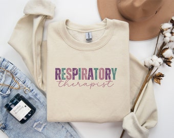 May include: A beige sweatshirt with the text "RESPIRATORY therapist" in colorful letters. The sweatshirt is laid out on a white surface with a pair of jeans, a candle, and a hat.
