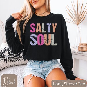 May include: Black long sleeve tee with the words "SALTY SOUL" in colorful, distressed lettering. The shirt is a crew neck style. The image also shows the words "Black" and "Long Sleeve Tee".