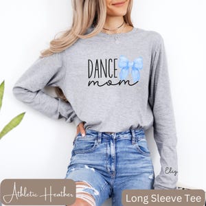 Może przedstawiać: Szara koszulka z długim rękawem z napisem "Dance Mom" i niebieską akwarelową kokardką. Koszulka wykonana jest z tkaniny athletic heather.
