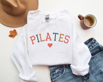 May include: White crewneck sweatshirt with the word "PILATES" in colorful letters and a red heart. The sweatshirt is paired with denim jeans, a brown hat, and a mug of coffee.