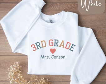 May include: A white long-sleeve crewneck sweatshirt featuring the text 3RD GRADE in arched, multi-colored letters above a small red heart and the name Mrs. Carson printed in dark teal. The word White is displayed in a rounded corner box.