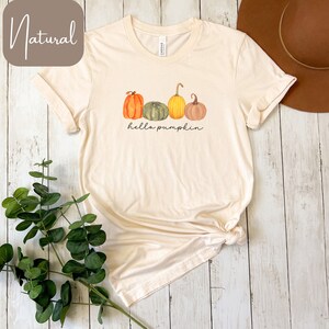 May include: Cream-colored t-shirt featuring a watercolor design of four pumpkins in shades of orange, green, yellow, and brown. The phrase "hello pumpkin" is written in a script font below the pumpkins. The shirt is folded and tied at the bottom.
