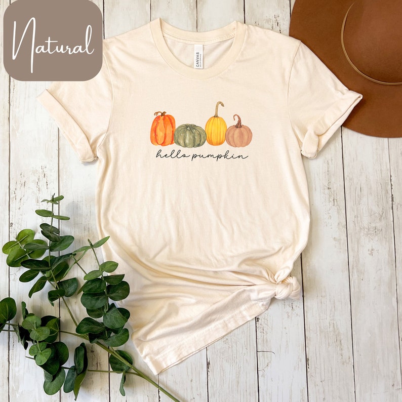 May include: Cream-colored t-shirt featuring a watercolor design of four pumpkins in shades of orange, green, yellow, and brown. The phrase "hello pumpkin" is written in a script font below the pumpkins. The shirt is folded and tied at the bottom.