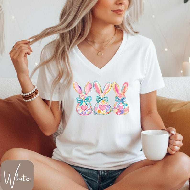 Op de afbeelding: Wit V-hals T-shirt met een aquarelontwerp van drie paashazen. Elke haas heeft een strik, een hart en kleurrijke bloempatronen. Een comfortabele, casual stijl.