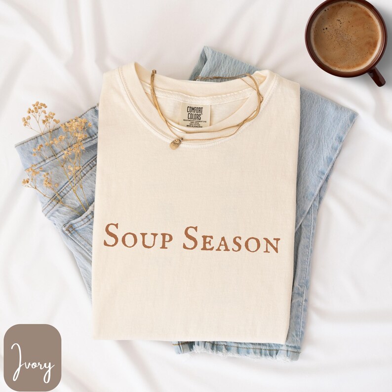 Puede incluir: Camiseta de color marfil con la frase "Soup Season" impresa en marr&oacute;n. La camiseta est&aacute; doblada y colocada sobre unos vaqueros azul claro. Tambi&eacute;n se ven un collar dorado y una taza de caf&eacute;.