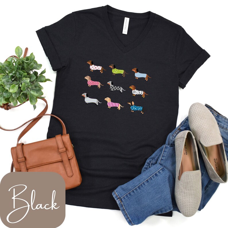May include: Black V-neck t-shirt with a pattern of dachshunds in sweaters. The dachshunds are in various colours and patterns. The shirt is styled with blue jeans, grey loafers, and a brown handbag.