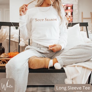 Puede incluir: Camiseta blanca de manga larga con el texto "Soup Season" en fuente marr&oacute;n. La camiseta se combina con pantalones de ch&aacute;ndal grises. La imagen tambi&eacute;n muestra una manta blanca y una calabaza decorativa.