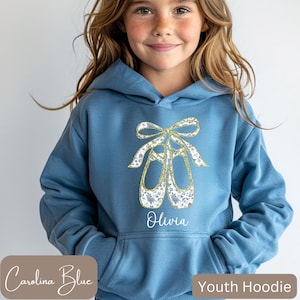 May include: Carolina blue youth hoodie featuring a gold and floral design of ballet shoes. The name "Olivia" is printed below the design. The hoodie has a drawstring hood and a front pocket. The text "Youth Hoodie" is printed on the bottom right.