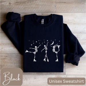 Puede incluir: Sudadera unisex negra con un gráfico blanco de esqueletos patinando sobre hielo bajo una luna creciente y estrellas. La sudadera tiene cuello redondo y mangas largas. La palabra "Black" está en la esquina inferior izquierda.