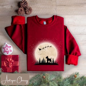 May include: A burgundy crewneck sweatshirt featuring a graphic of Santa Claus in a sleigh, a dog, and a full moon. The design is in black and white. The sweatshirt is surrounded by Christmas decorations.