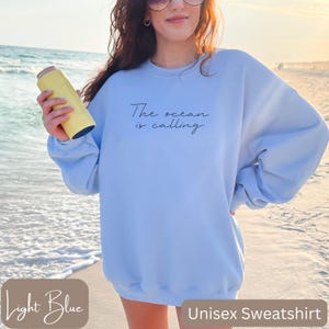 K&ouml;nnte beinhalten: Hellblauer Unisex-Sweatshirt mit dem Text "The ocean is calling" in Schwarz.