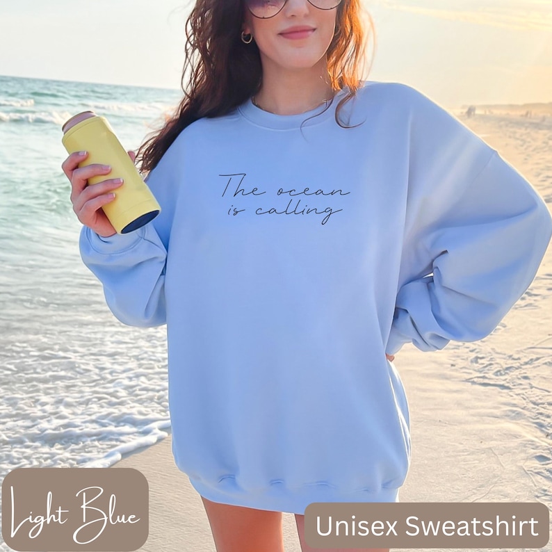 K&ouml;nnte beinhalten: Hellblauer Unisex-Sweatshirt mit dem Text "The ocean is calling" in Schwarz.
