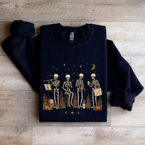 Puede incluir: Una sudadera de cuello redondo en color azul marino oscuro con un estampado gráfico de cuatro esqueletos realizando diversas actividades, como tocar la flauta, tejer y sostener una taza de café, rodeados de calabazas naranjas y flores de otoño.
