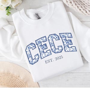 May include: White sweatshirt with the name "CECE" in blue floral print lettering. The text "EST. 2025" is below the name. The sweatshirt is displayed on a light-colored surface with floral and woven accents.